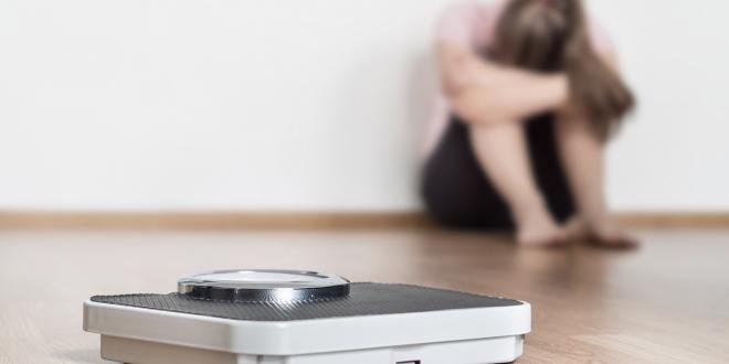 A scale on the floor with a woman sitting in the background with her head hung with failure.